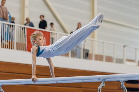 Focused young boy performs a pike hold on parallel bars, legs raised high, showcasing strength and concentration at a gymnastics meet.