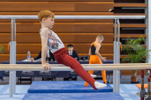 Red-haired boy sits intently on parallel bars, legs extended, focused expression during a gymnastics meet.