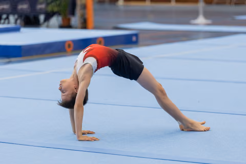 Young gymnast performs a bridge pose on blue floor mat, head inverted, body arched with focused control.