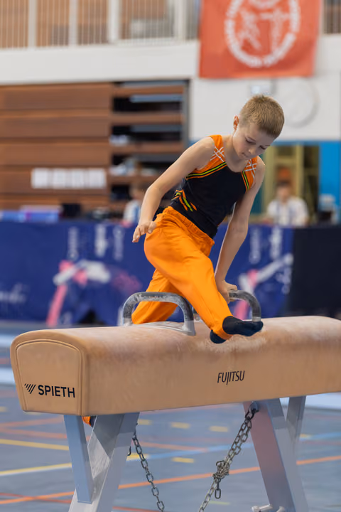 Focused young boy in orange pants performs on a Spieth pommel horse during a gymnastics competition.