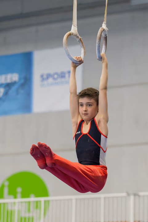 Young male gymnast hangs from still rings in a pike position, wearing red and black, with intense focus on his face.