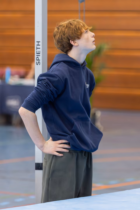 Red-haired teen gymnast looks upward with hands on hips, focused and composed beside a Spieth apparatus in a gym.