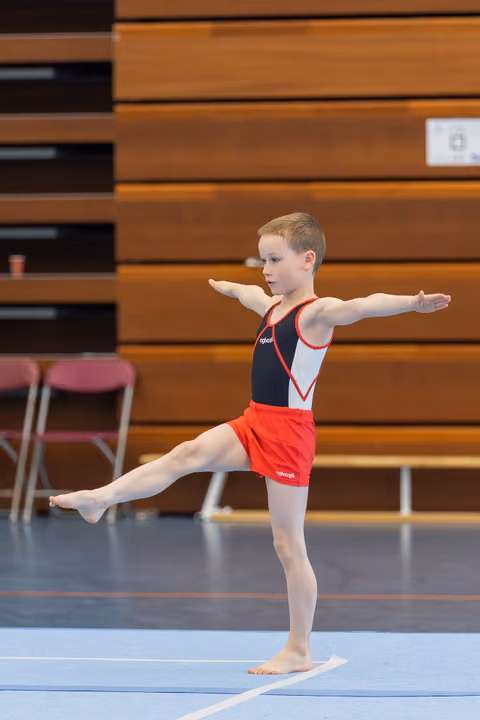 Young boy gymnast balances on one leg with arms outstretched, focused and composed during a floor routine in a sports hall.