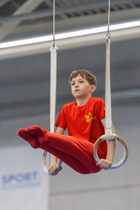 Young boy in red grips gymnastics rings mid-routine, legs raised, expression calm and concentrated in a sports hall.