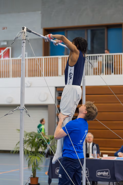 Coach lifts gymnast to reach the high bar in a busy indoor gymnastics hall, focused and determined.