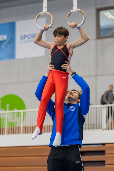 Young male gymnast grips still rings with focused expression while coach in blue jacket steadies him at the waist.