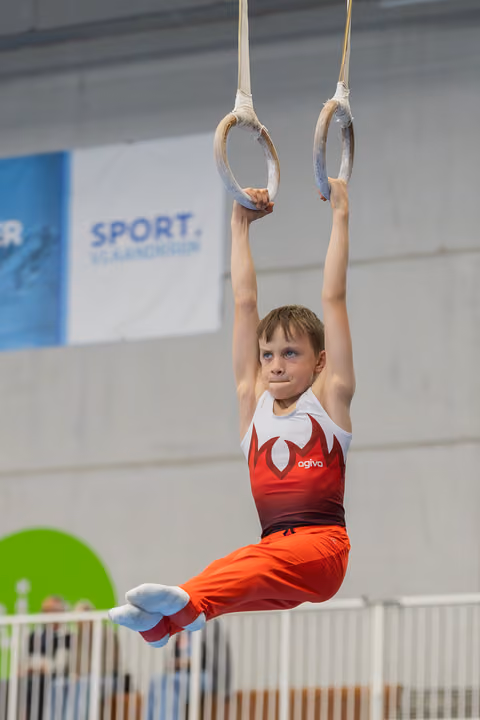 Young male gymnast hangs from still rings in a tuck position, focused expression, wearing red and white agiva leotard.