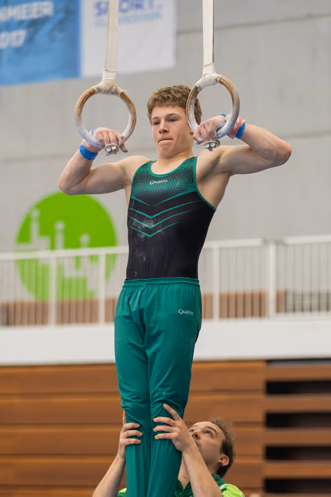 Young male gymnast grips still rings with intense focus, supported from below by a coach in a gym.