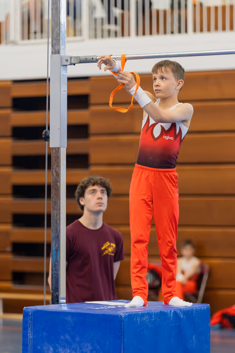 Young boy in red leotard grips the horizontal bar with focused concentration while a coach watches from below.