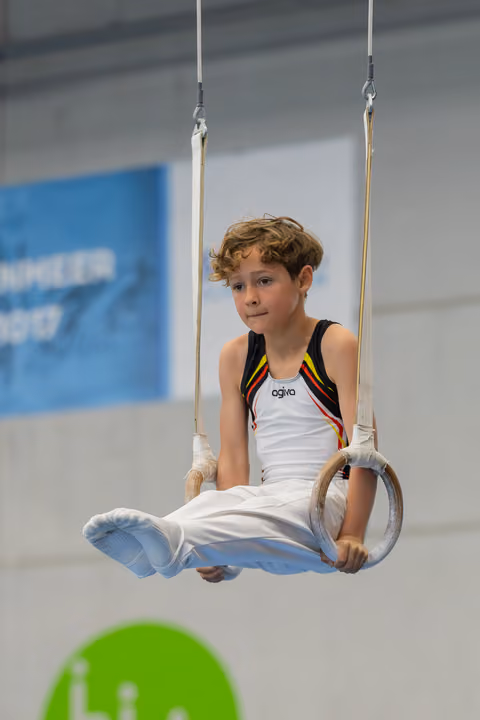 Young boy sits calmly on gymnastics rings, wearing a Belgian leotard, with a composed, attentive expression in an indoor gym.