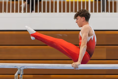 Young male gymnast performs on parallel bars, legs raised with intense focus, wearing red Canadian-style leotard in gymnasium.