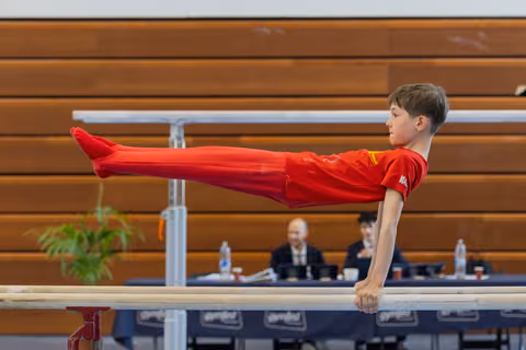 Focused young boy in red holds a perfect straddle-L on parallel bars before judges at a gymnastics meet.