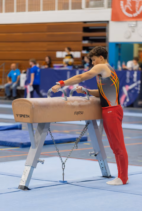 Young male gymnast grips pommel horse handles, focused and composed, preparing his routine in a busy gymnasium.