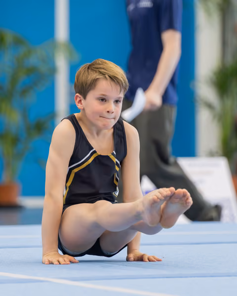 Young boy gymnast seated on blue mat, legs raised, looking focused and composed during a gymnastics event.