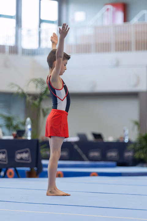 Young male gymnast stands tall on the floor mat, one arm raised in a confident salute before his routine begins.