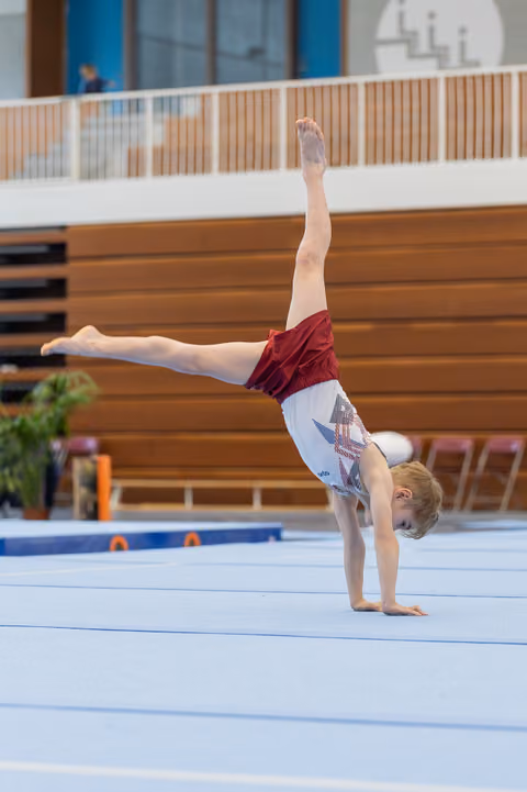 Young male gymnast performs a one-armed handstand with legs split on a blue gymnastics floor mat indoors.