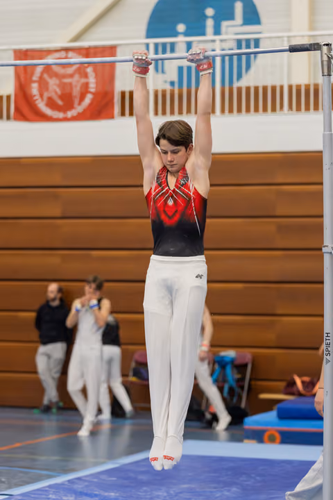 Young male gymnast hangs from the high bar with arms extended, wearing a red and black leotard, focused expression.
