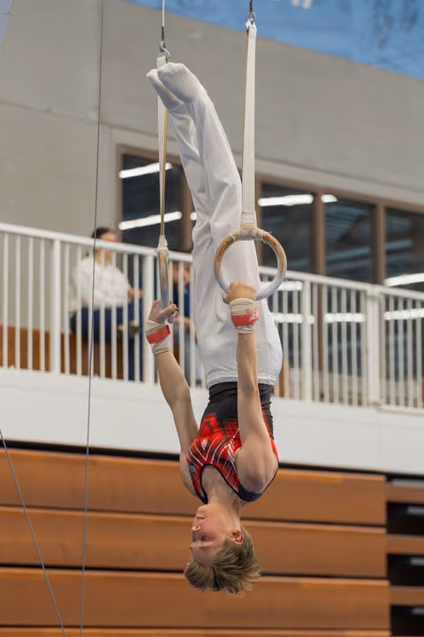 Young male gymnast hangs inverted on still rings, smiling upside-down, wearing red plaid shorts in an indoor gymnasium.