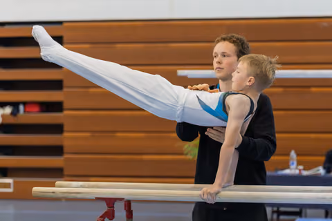 A teen coach spots a young boy performing a horizontal hold on parallel bars, both focused and composed in a gymnasium.