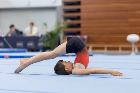 Male gymnast performs a dramatic backbend arch on the floor mat, legs extended overhead, in a competitive gymnastics hall.