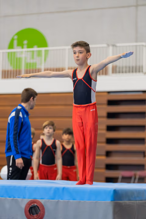 Young male gymnast stands focused on apparatus, arms outstretched for balance, as teammates and coach watch in background.