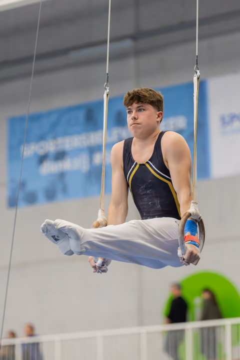 Young male gymnast holds a tuck position on still rings, wearing a navy leotard, with a focused, determined expression.