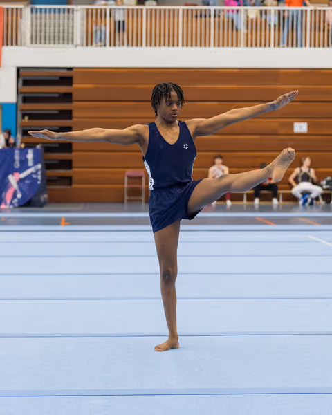 Young male gymnast holds a precise arabesque on one leg, arms extended, focused expression on a blue competition floor.