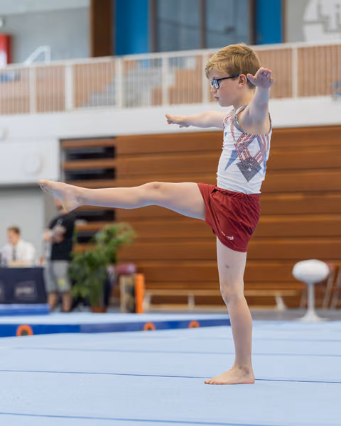 Young boy with glasses performs a precise leg kick on the floor exercise mat, arms extended, expression focused and determined.