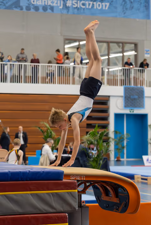 Young male gymnast performs a handstand on the vault, legs straight up, concentrated expression in a busy sports hall.