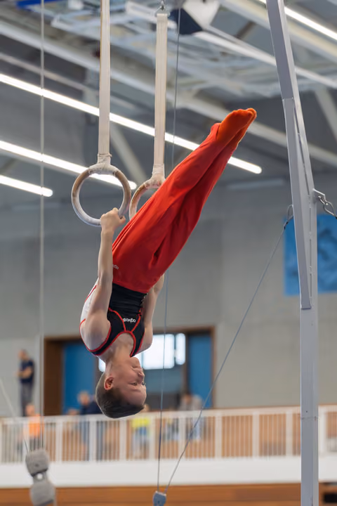 Young male gymnast hangs inverted on still rings, legs raised, wearing red and black, performing in an indoor gymnasium.