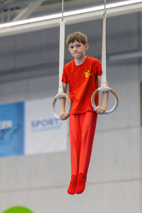Young boy in red gymnastics suit holds still rings, expression calm and concentrated in an indoor sports hall.