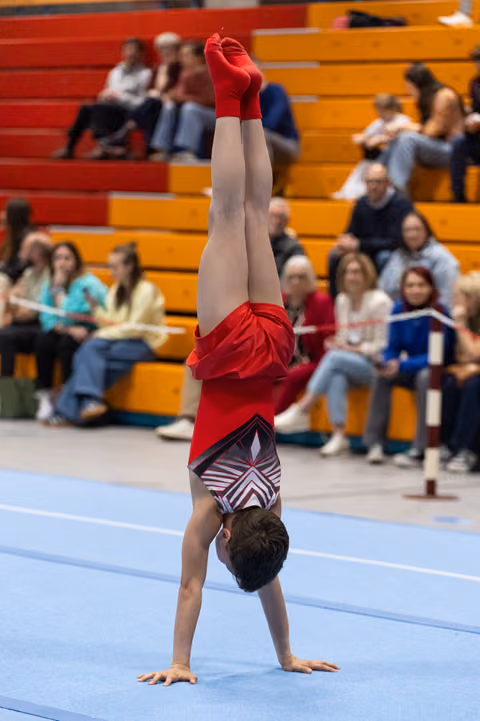 Young gymnast performs a handstand on floor exercise mat wearing red warmup jacket, with spectators in bleachers