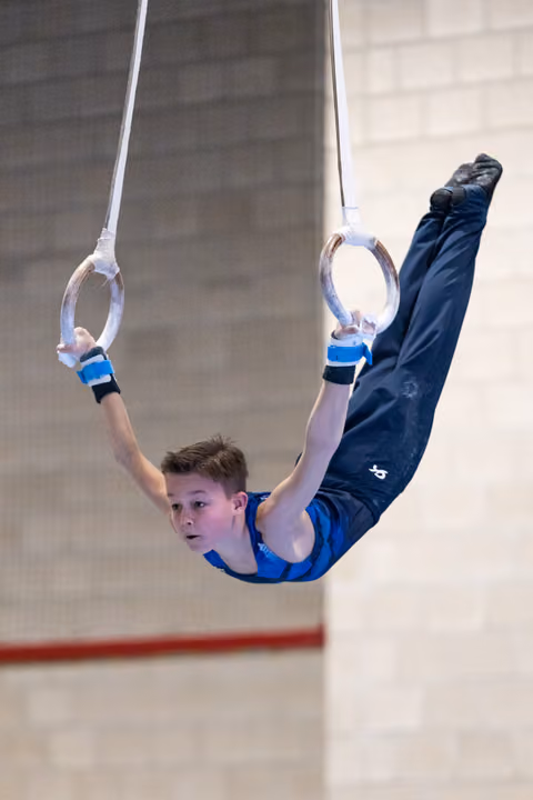 Young male gymnast performs an inverted pike position on rings, displaying strength and control in blue competition attire