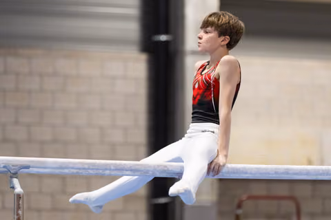 Young gymnast in red and black leotard sits on parallel bars with focused expression during routine