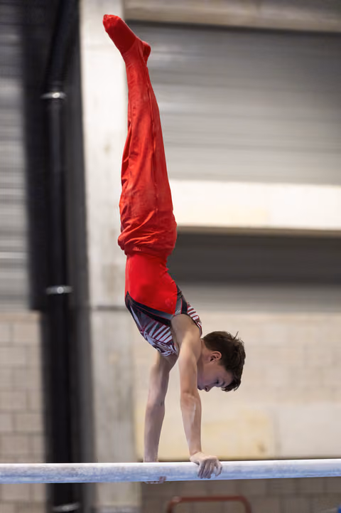 Male gymnast performing a handstand on the balance beam, legs extended vertically in red pants during training