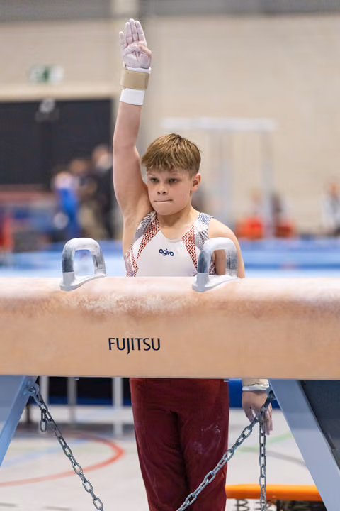 Young gymnast raises hand in salute while gripping pommel horse handles, displaying focus and determination during routine