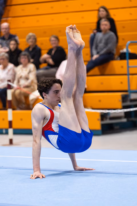 Male gymnast performs an L-support position during floor exercise routine, demonstrating strength and control before spectators