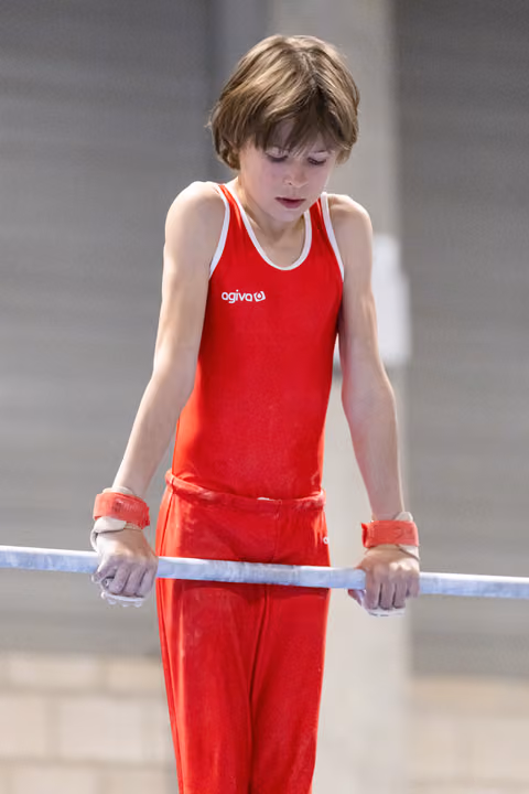 Young gymnast in red uniform focuses intently on the parallel bars, hands gripped and eyes downcast in concentration