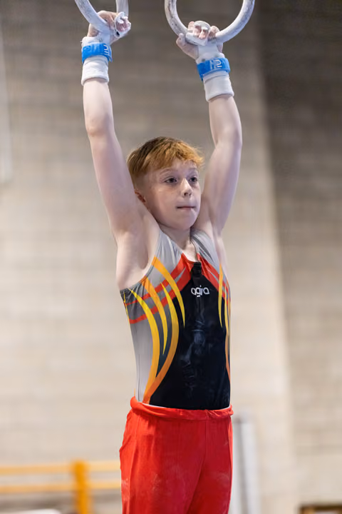 Young redhead gymnast hanging from rings with focused expression, wearing colorful leotard and red shorts in training hall