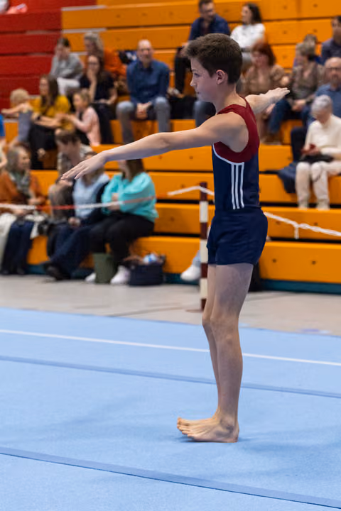 Young male gymnast in navy and maroon leotard stands barefoot in ready position on floor mat, arms extended, spectators watching