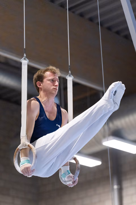 Male gymnast performs on rings with extended body position, wearing navy leotard and white pants in indoor facility