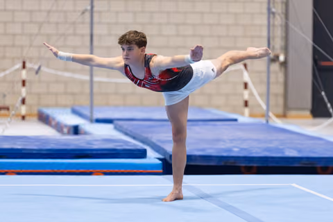 Young gymnast performs an arabesque balance on floor exercise, arms extended gracefully in a gymnastics training facility