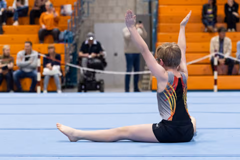 Young gymnast performs a straddle split with raised arms during floor routine at indoor gymnasium meet
