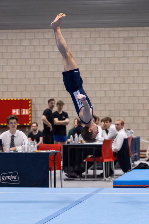 Male gymnast performs inverted backflip on floor exercise while judges observe from tables in indoor venue