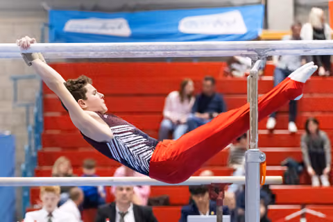 Male gymnast performs horizontal bar routine with extended body position over red bleachers filled with spectators