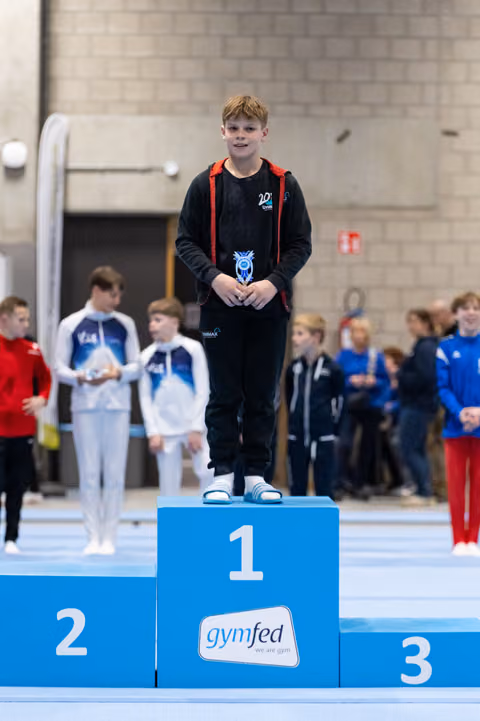 Young gymnast in black tracksuit stands proudly on first place podium holding medal, with competitors visible behind