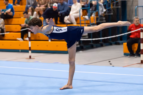 Young gymnast performs an elegant arabesque balance during floor routine at indoor meet with spectators in bleachers