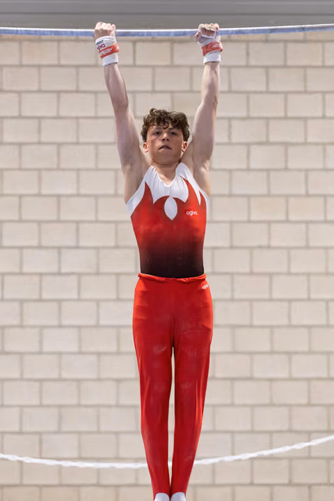 Young male gymnast hangs from horizontal bar in red and white leotard, arms extended overhead, focused expression
