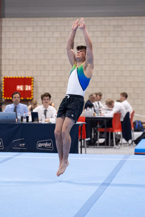 Male gymnast performing a vertical jump with arms raised overhead during floor exercise routine at indoor competition