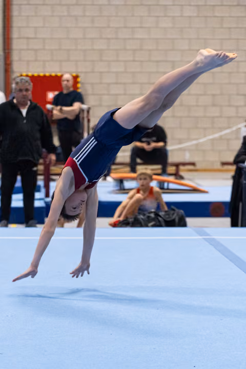 Young gymnast performs inverted handstand during floor routine while coaches and teammates watch in gym background